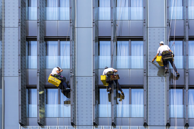 Three workers with harnesses and cleaning equipment cleaning a building's facade